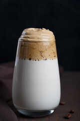Single glass of homemade popular korean dalgona drink made by whipping instant coffee powder, sugar and hot water with addition of cold milk on dark wooden background at kitchen. Vertical orientation