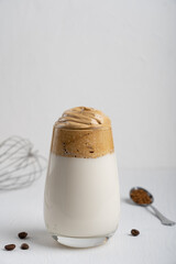 Single glass of popular korean dalgona drink made by whipping instant coffee powder, sugar and hot water with addition of cold milk on white wooden background with kitchen utensil. Vrtical orientation