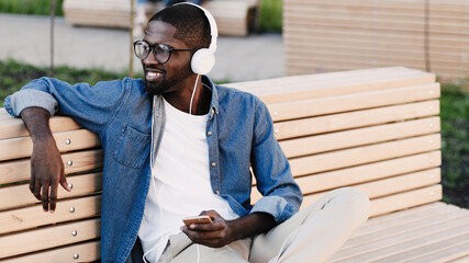 Stylish guy in denim shirt and white headphones listening music while sitting on bench in park. Young African American man is listening music outdoors and smiling.