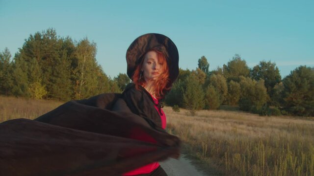 Pretty carefree redhead sorceress in conical hat, gracefully whirling with black cloack fluttering in the wind in rays of beautiful sunset while walking on dirt road on halloween over natural