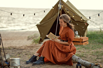A beautiful girl in a brown dress reads a book against the background of a tent.