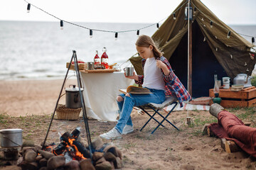 A girl in a plaid shirt reads a book next to a campfire against the background of a lake.