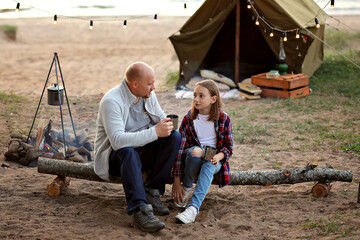 Father and daughter sit on a log against the background of a tent and a lake.