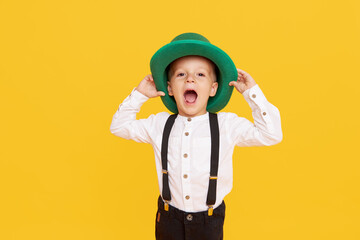 Smiling little child boy in green leprechaun hat on yellow background. St. Patrick Day celebration. Funny face