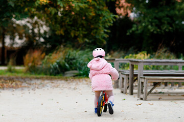 Fototapeta premium Cute little toddler girl with helmet riding on run balance bike to daycare, playschool or kindergarden. Happy child having fun with learning on learner bicycle. Active kid on cold autumn day outdoors.