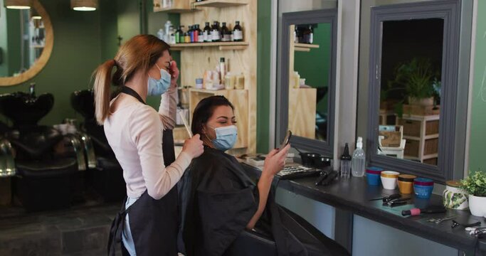 Female hairdresser and female customer wearing face masks looking at smartphone at hair salon