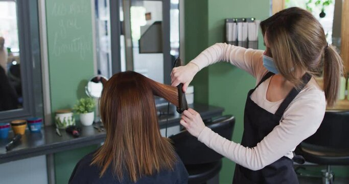 Female hairdresser wearing face mask straightening hair of female customer at hair salon