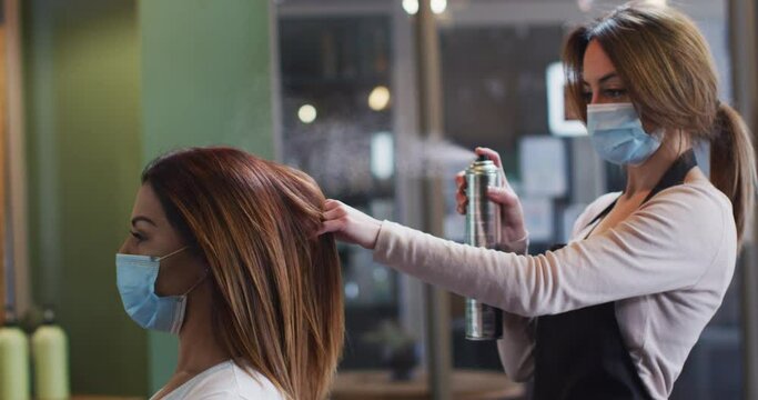 Female hairdresser wearing face mask spraying hairspray on hair of female customer at hair salon