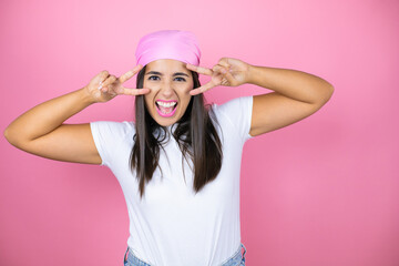 Young beautiful woman wearing pink headscarf over isolated pink background Doing peace symbol with fingers over face, smiling cheerful showing victory