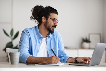 Indian male entrepreneur using laptop and taking notes while working in office