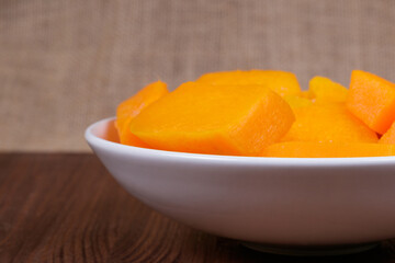 Pieces of pumpkin in the bowl at the wooden table and sackcloth background.