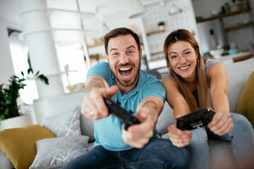 Obraz premium Husband and wife playing video game with joysticks in living room. Loving couple are playing video games at home.