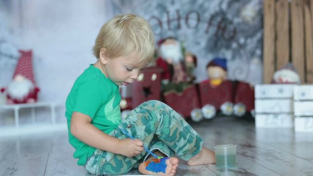 Cute blond toddler child, boy, painting his feet with colorful paints at home