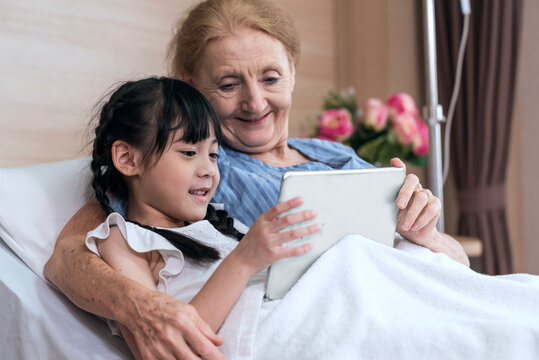 Asian Lovely girl and grandmother watching fun and happy video together on tablet on patient bed. granddaughter visit the sick grandmother to encourage in hospital - Powered by Adobe