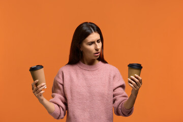 Photo of confused unaware woman in pink sweater chooses between two types of drink in disposable...