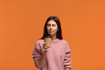 Wonderful aroma. Photo of satisfied woman in pink sweater holds paper cup of hot drink, closes eyes. Beautiful young woman enjoys aromatic tea or coffee. Isolated on orange wall