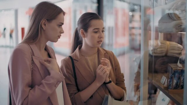 Medium shot of happy young female friends standing before store window in mall and chatting