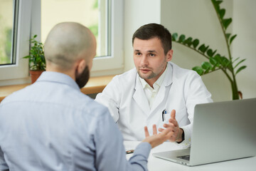 A caucasian doctor in a white lab coat is sitting at a desk and describing the treatment of a bald...