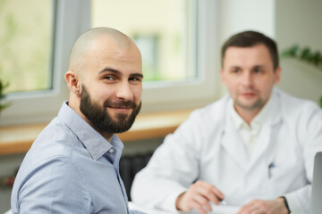 Obraz premium A bald male patient is smiling near a caucasian doctor in a white lab coat in a hospital. A man with a beard at an appointment in a doctor's office.