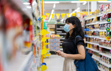 Asian women are shopping at the grocery store, holding bphone and wearing a health mask to prevent infection. She's picking goods from the shelves in lifestyle shopping concept,Soft Focus.