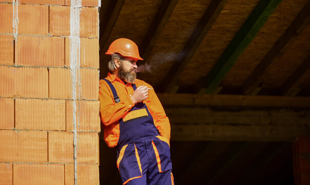construction worker smoking cigarette. building is under construction. bearded man in uniform and hard hat. copy space. repairman engineer wear helmet on site. tired builder relax. time to relax