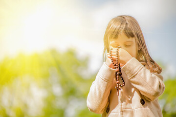 Little child girl praying with a sweet expression. She looks down and holding a wooden rosary.