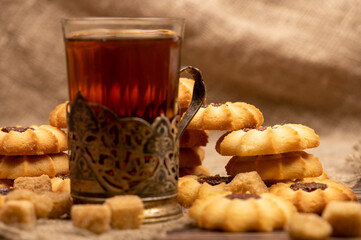 Homemade pastry cookies with jam, a faceted glass of tea in a vintage Cup holder and pieces of brown cane sugar on a background of homespun fabric with a rough texture, close-up, selective focus.