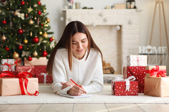 Young Beautiful Woman Writing Shopping Or Wish List For Christmas In Festive Interior At Home