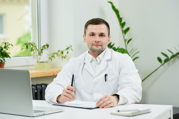 A caucasian doctor in a white lab coat is posing near a laptop in a hospital. A male therapist is...