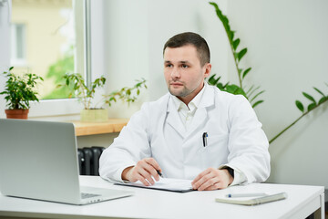 A doctor in a white lab coat is listening to a patient on an online meeting on a laptop in a hospital. A therapist is doing his work on a video call on a computer.  An online doctor's appointment.