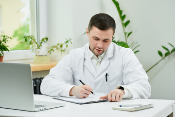 Fototapeta premium A caucasian doctor in a white lab coat is filling out a patient card with a diagnosis near a laptop in a hospital. A therapist is waiting for a patient in a doctor's office.