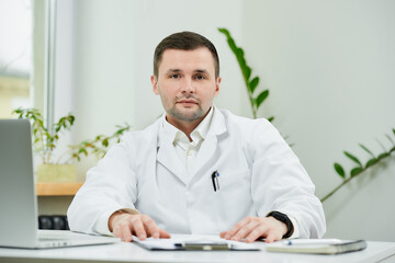 Fototapeta premium A kind caucasian doctor in a white lab coat is sitting relaxed at his desk and listening intently in a hospital. A therapist near a laptop is waiting for a patient in a doctor's office.