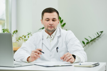 Fototapeta premium A caucasian doctor in a white lab coat is sitting at his desk and lowered his eyes during examining the patient’s medical history in a hospital. A therapist near a laptop in a doctor's office.