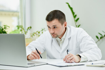A serious caucasian doctor in a white lab coat is carefully entering the patient’s medical...