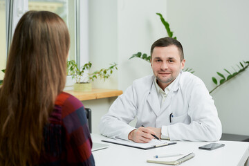 A caucasian doctor in a white lab coat is sitting at a desk and listening to a female patient's...