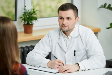 A caucasian doctor in a white lab coat is sitting at a desk near a female patient in a doctor's office. A woman with long hair at a doctor's appointment in a hospital.