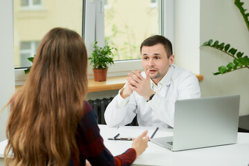 A caucasian doctor in a white lab coat is sitting at a desk and discussing the treatment with a female patient in a doctor's office. A woman with long hair at a doctor's appointment in a hospital.