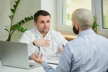 Naklejka na ściany i meble A caucasian doctor in a white lab coat is sitting at a desk and arguing about the treatment of a bald male patient in a hospital. A man with a beard at an appointment in a doctor's office.