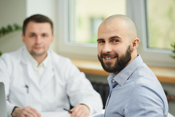A bald male patient is smiling near a caucasian doctor in a white lab coat in a hospital. A happy man with a beard at an appointment in a doctor's office.
