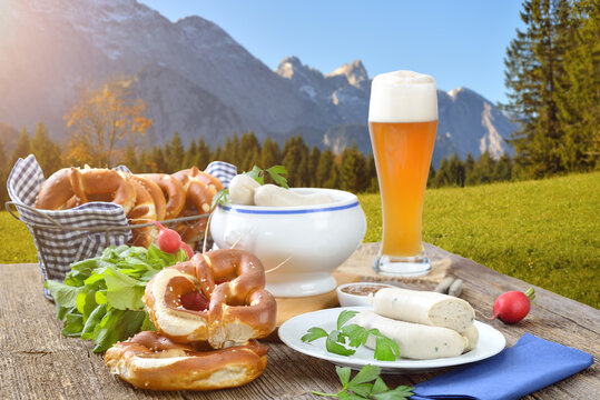 Deftiges, bayerisches Weißwurstfrühstück in den Bergen – Hearty Bavarian breakfast with veal sausages, fresh pretzels and a wheat beer outside in the nature near Garmisch-Partenkirchen