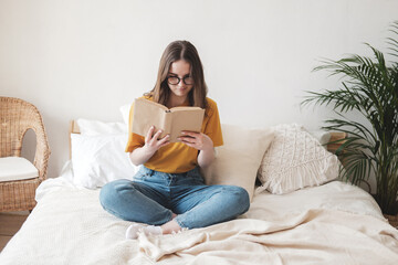 Obraz na płótnie Canvas Young beautiful girl student in glasses, an orange T-shirt and blue jeans sits on bed with pillows and reads paper book. Self-education concept at home during quarantine. Home schooling, hobby.