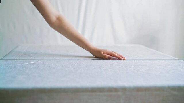 CU: young woman makes marks on white linen fabric sheet with chalk and long metal ruler working at table in light room close view