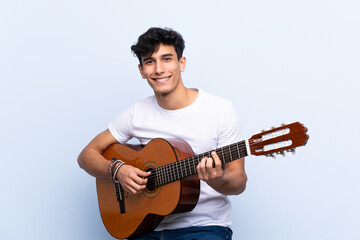 Obraz premium Young Argentinian man with guitar over isolated blue background smiling a lot
