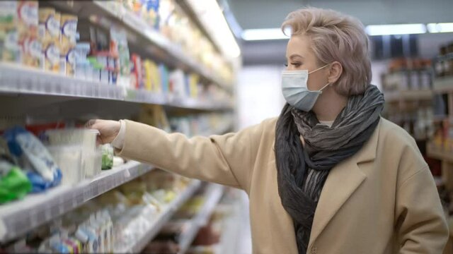 Young woman volunteer in a medical protective mask picks cottage cheese while in a supermarket during a pandemic. Shopping, quarantine, coronavirus. Health, customer care.