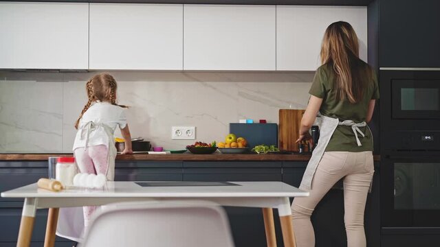 young mother with adorable little daughter in aprons cooks delicious dinner together in light modern kitchen at home backside view