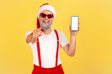 Extremely happy cheerful grey bearded man in santa claus costume pointing finger at camera showing smartphone with empty display, holidays bonuses. Indoor studio shot isolated on yellow background