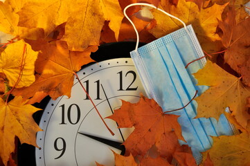 Wall clock and medical mask on the background of yellow autumn leaves.