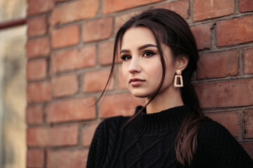 Close up lifestyle portrait of young indian woman standing on the street cloudy weather, leaning against the brick wall, dressed in black pullover and looking away. 