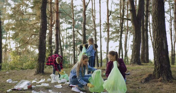Beautiful satisfied active female volunteers picking up rubbish into the plastic bags from park territory and giving high five each other on the background of another working members of their team