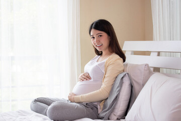 Obraz premium Young beautiful pregnant asian woman sitting on bed and touching belly near the window in bedroom. Pregnancy, motherhood, people and expectation concept.